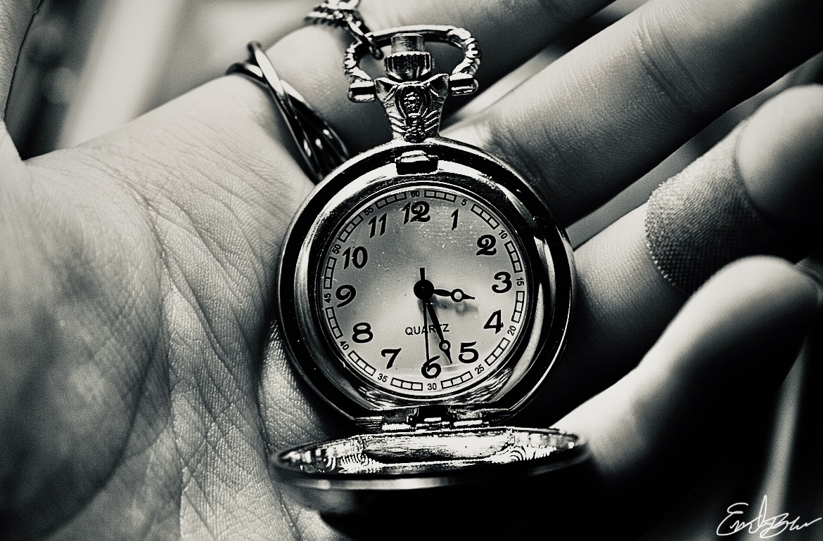 A black and white depiction of a hand holding “time” an open pocket watch 