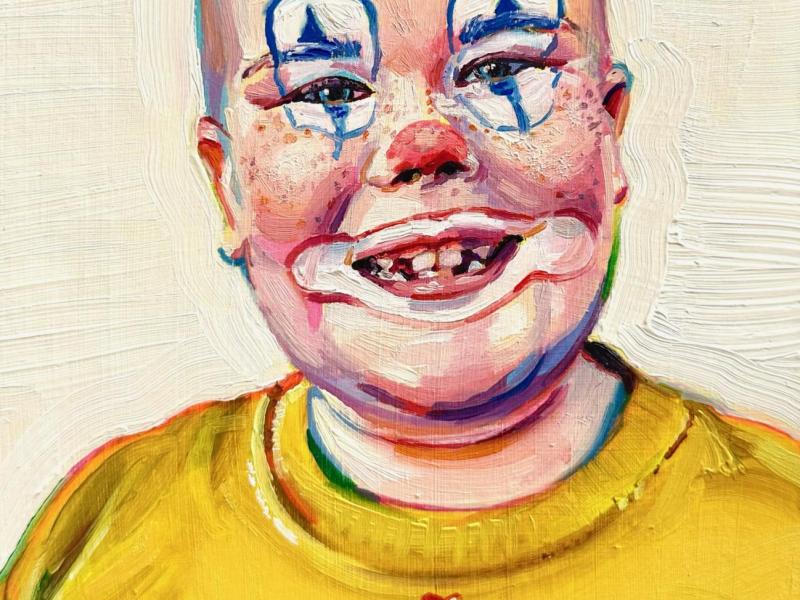 A young boy in a yellow shirt with clown makeup. He's bald and is missing teeth. The paint is chunky and colorful. 