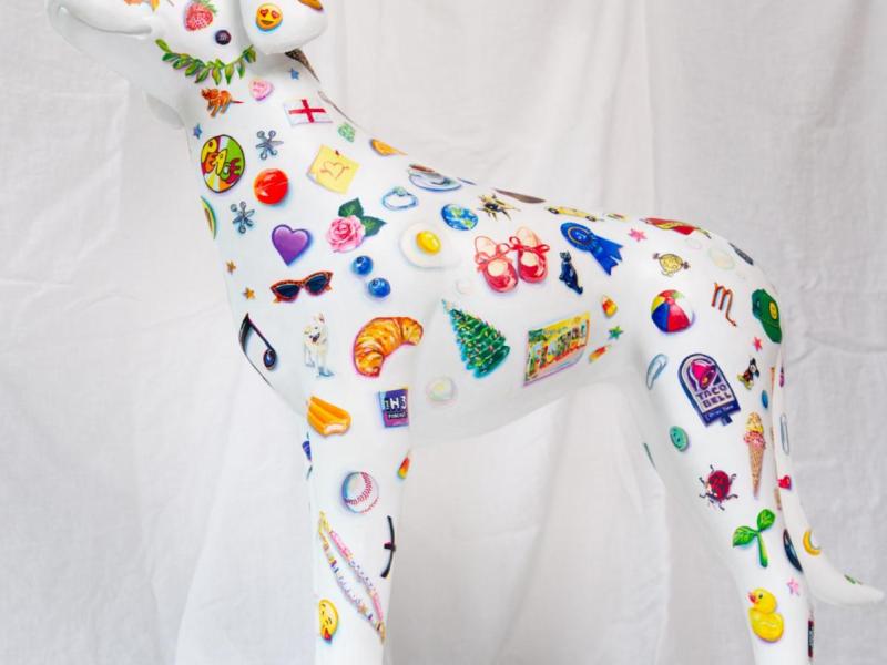 A white dog sculpture painted with dozens of tiny little objects, such as hearts, fruit, plants, logos, toys, etc. 