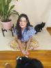 Artist Michelle Hawran sitting on the ground with her black cat smiling up at the camera. There is a plant behind her. 